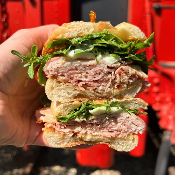A hand holds a sliced sandwich from The Depot, revealing layers of meat, arugula, and sauce.