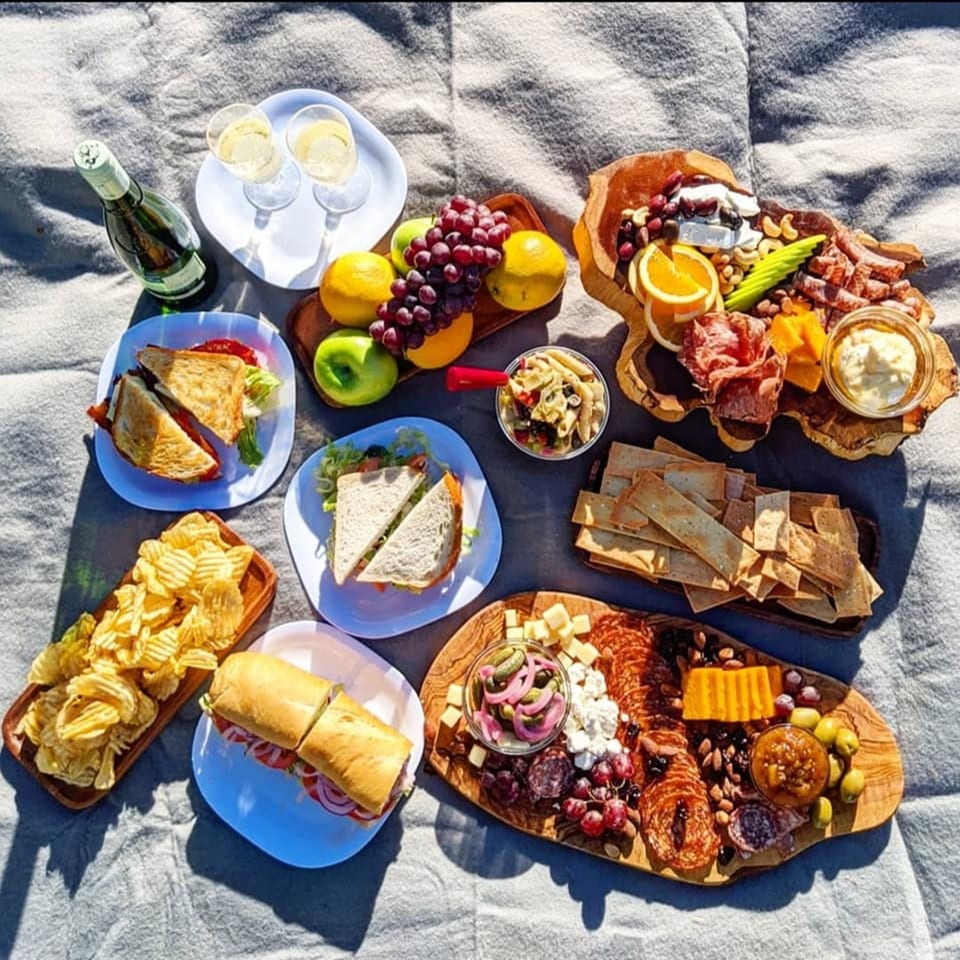 Overhead view of a picnic spread with sandwiches, charcuterie boards, chips, fruit, and champagne.