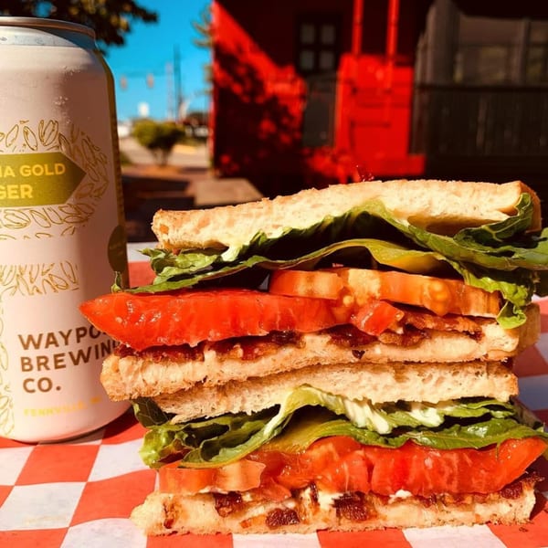 A stacked BLT sandwich with lettuce, tomato, and bacon, next to a beer can on a red and white checkered paper.