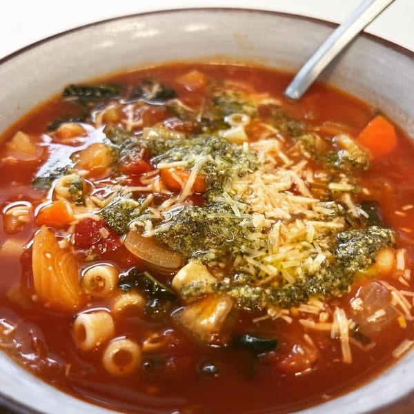 Close-up of a hearty minestrone soup with pasta, vegetables, pesto, and shredded cheese in a bowl.
