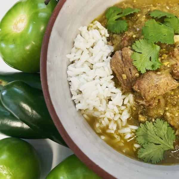 Close-up of a bowl of green chili stew with rice and cilantro, surrounded by fresh green peppers.
